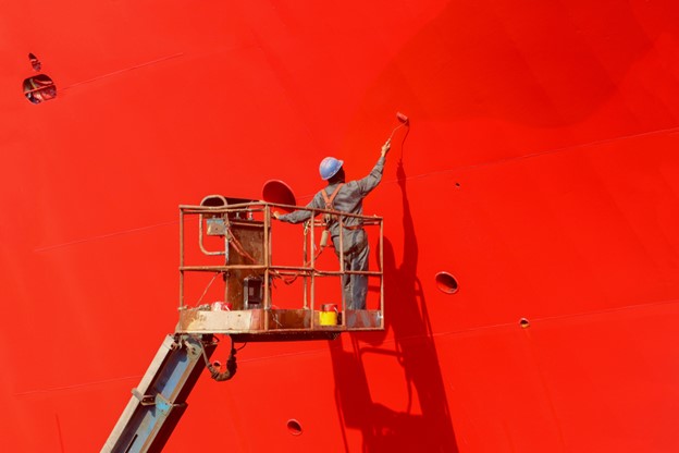 Worker painting ship hull on sherry picker using paint roller